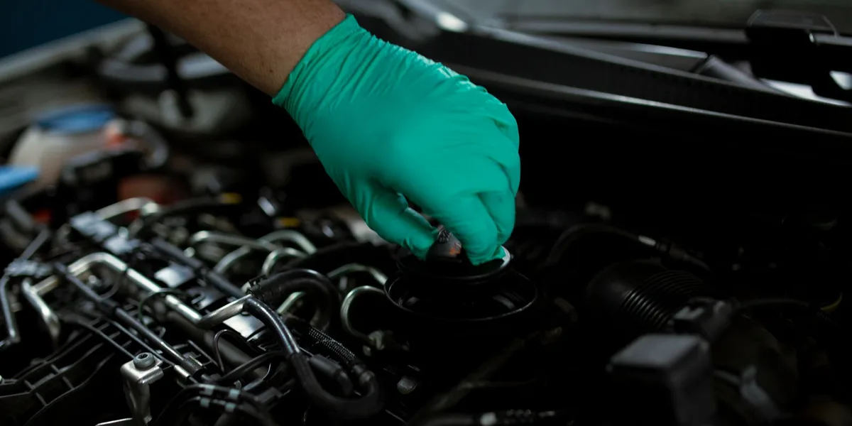A gloved hand lifting the oil filler cap off the cam cover of a modern car engine with the hood raised overhead