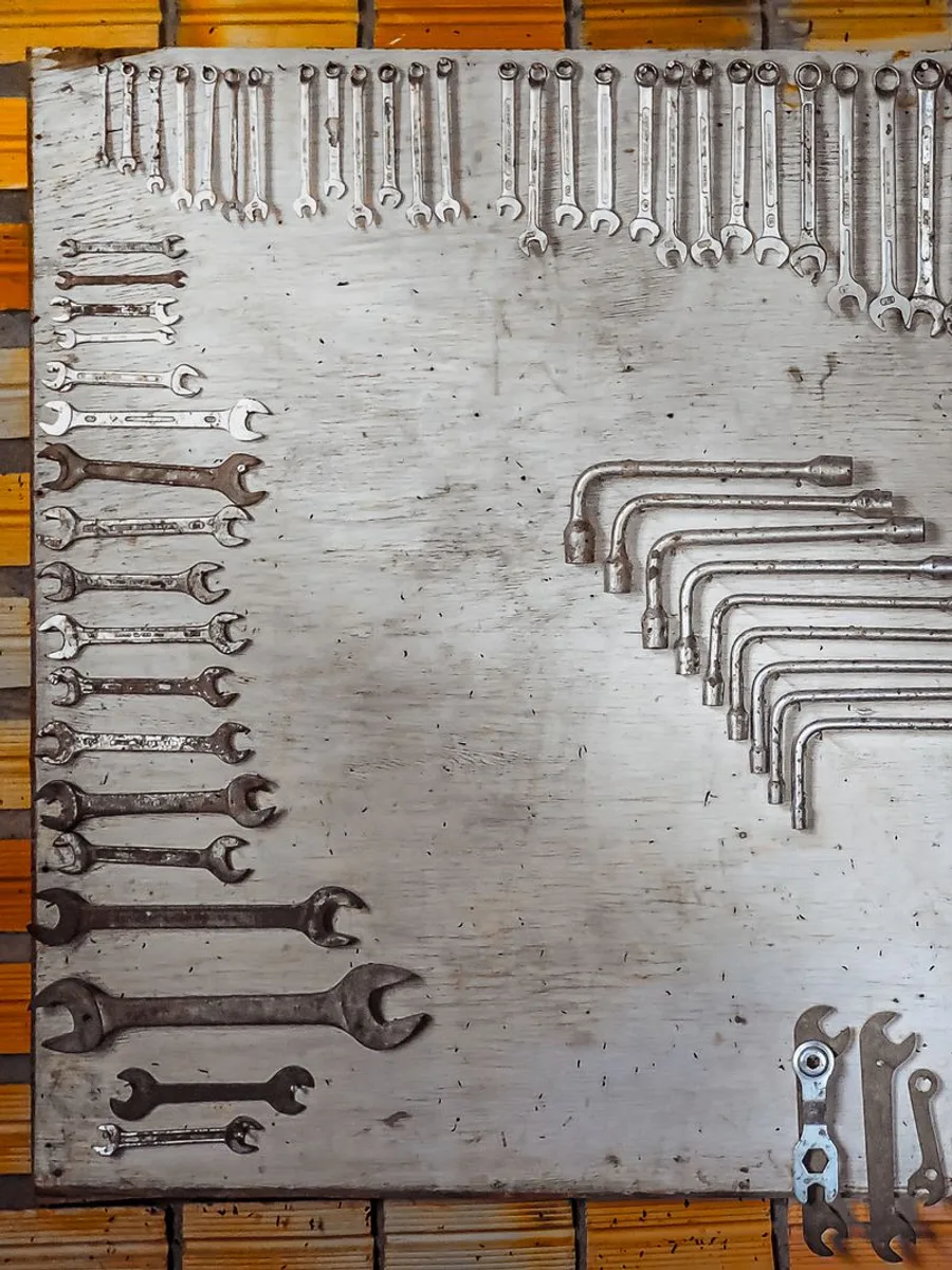 Rows of open-end and combination wrenches and L-shaped socket bars hung in size order on a whitewashed plywood tool board mounted to an amber slatted workshop wall